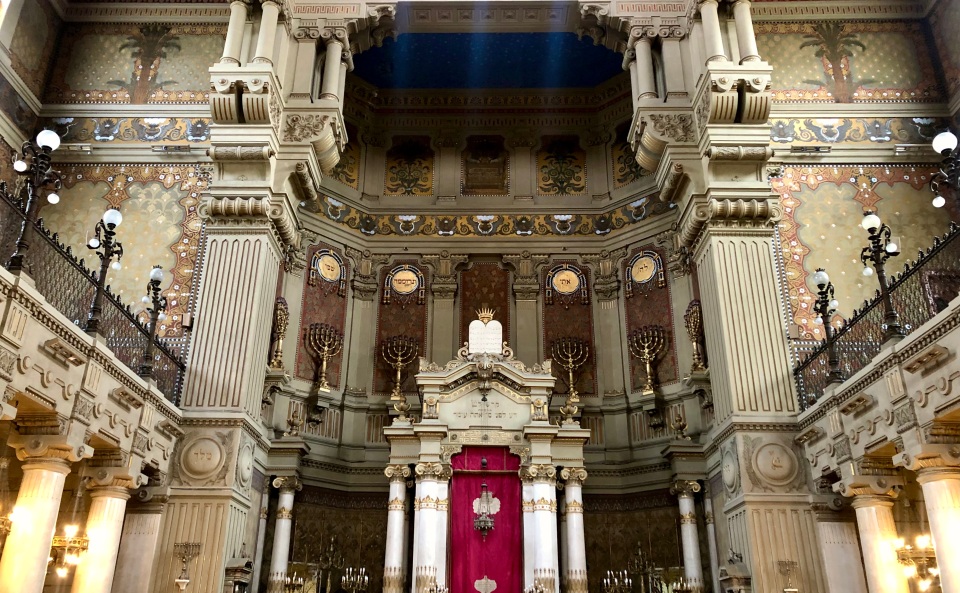The interior of the Great Synagogue of Rome, lavish with Greek columns, golden menorahs, and frescoes of palm trees and decorative patterns on the walls.