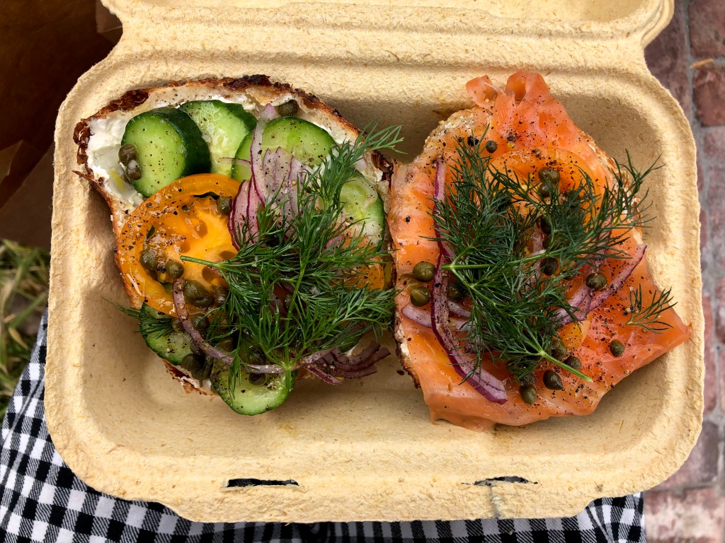 Two decadent bagel-halves sit in a to-go tray. One is loaded with fresh heirloom tomatoes and cucumbers, while the other is layered with a healthy serving of lox. Both are topped generously with fresh dill and capers.